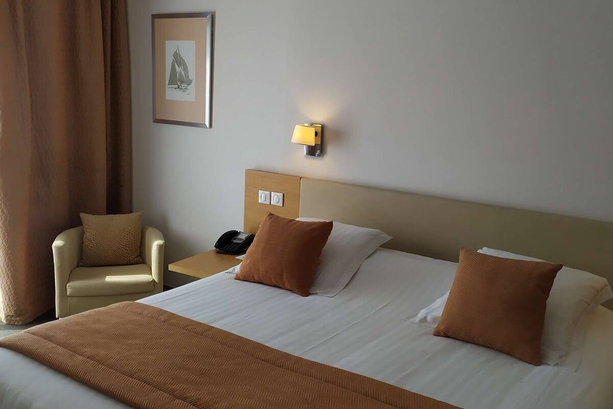 A hotel room with a large bed, two pillows, a chair, a bedside table with a phone, and a wall-mounted lamp.