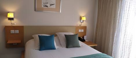 Habitación de hotel con una cama grande, dos lámparas de noche, un cuadro en la pared y una ventana con cortinas.