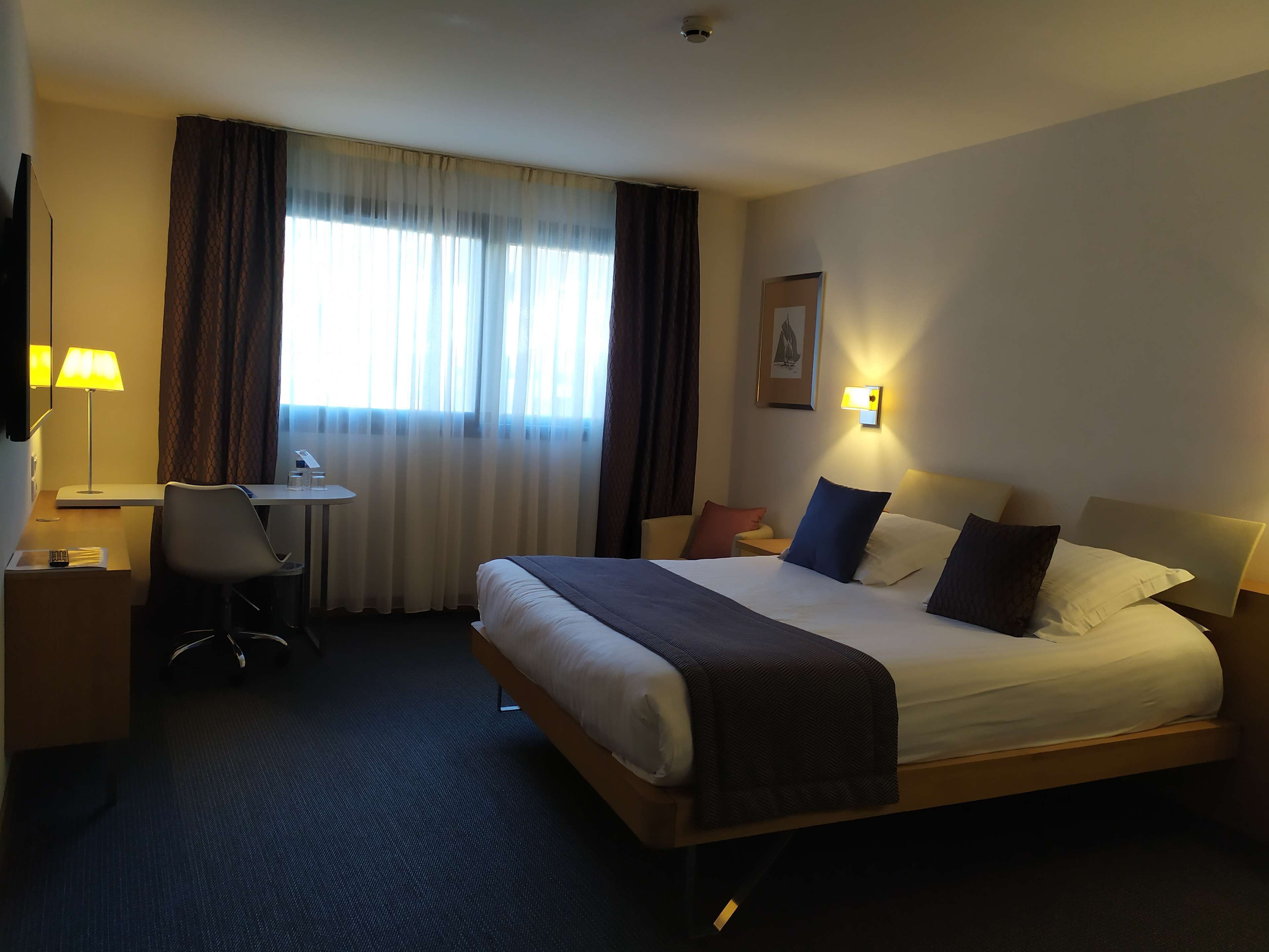 A hotel room with a large bed, a desk, a chair, and a lamp.