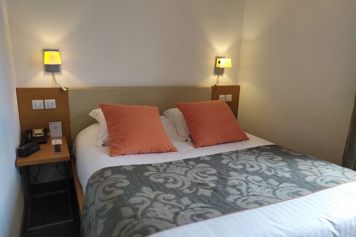A hotel room with a bed, two pillows, a bedside table with a phone and a lamp, and a wall-mounted light.