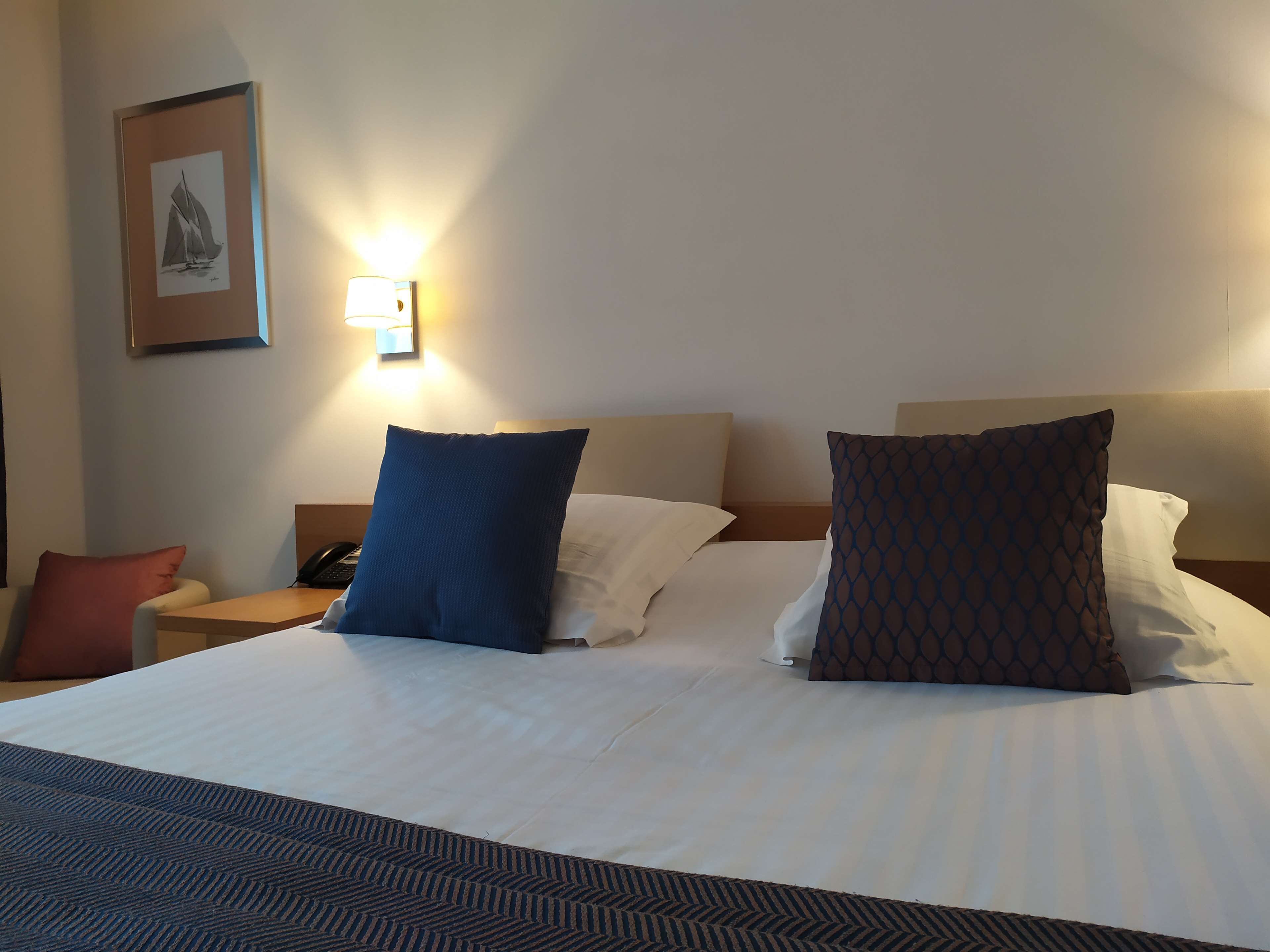 A neatly made bed with white linens and two pillows, a bedside table with a phone, and a wall-mounted light fixture.