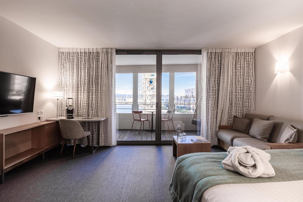 A hotel room with a bed, a desk, a TV, a balcony with a view of the city, and a sofa.