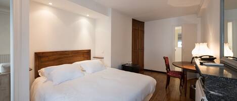 A hotel room with a large bed, a wooden headboard, a dining table with chairs, a small side table, and a mirror above the counter.