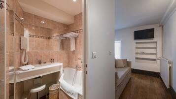 Une salle de bain moderne avec une baignoire, un lavabo et des toilettes. On y trouve un porte-serviettes, une poubelle et une étagère avec des articles de toilette. La pièce est dotée d’un plancher en bois et d’un mur blanc.