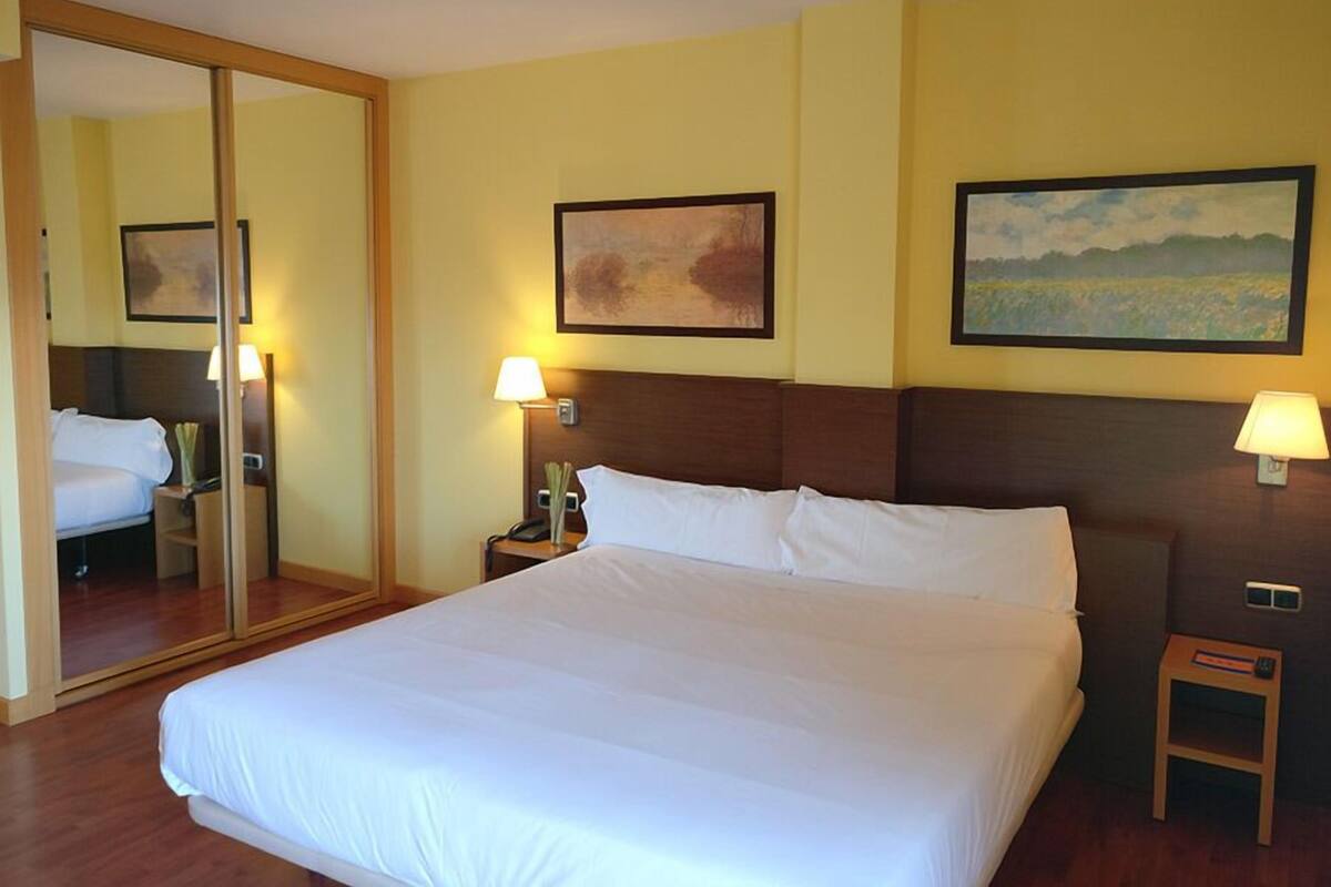 Habitación de hotel con una cama grande, dos mesitas de noche con lámparas, un espejo y dos obras de arte enmarcadas en las paredes.