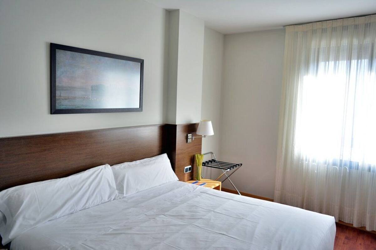 Habitación de hotel con una cama grande, un cuadro enmarcado en la pared, una lámpara de noche y una ventana con cortinas traslúcidas.