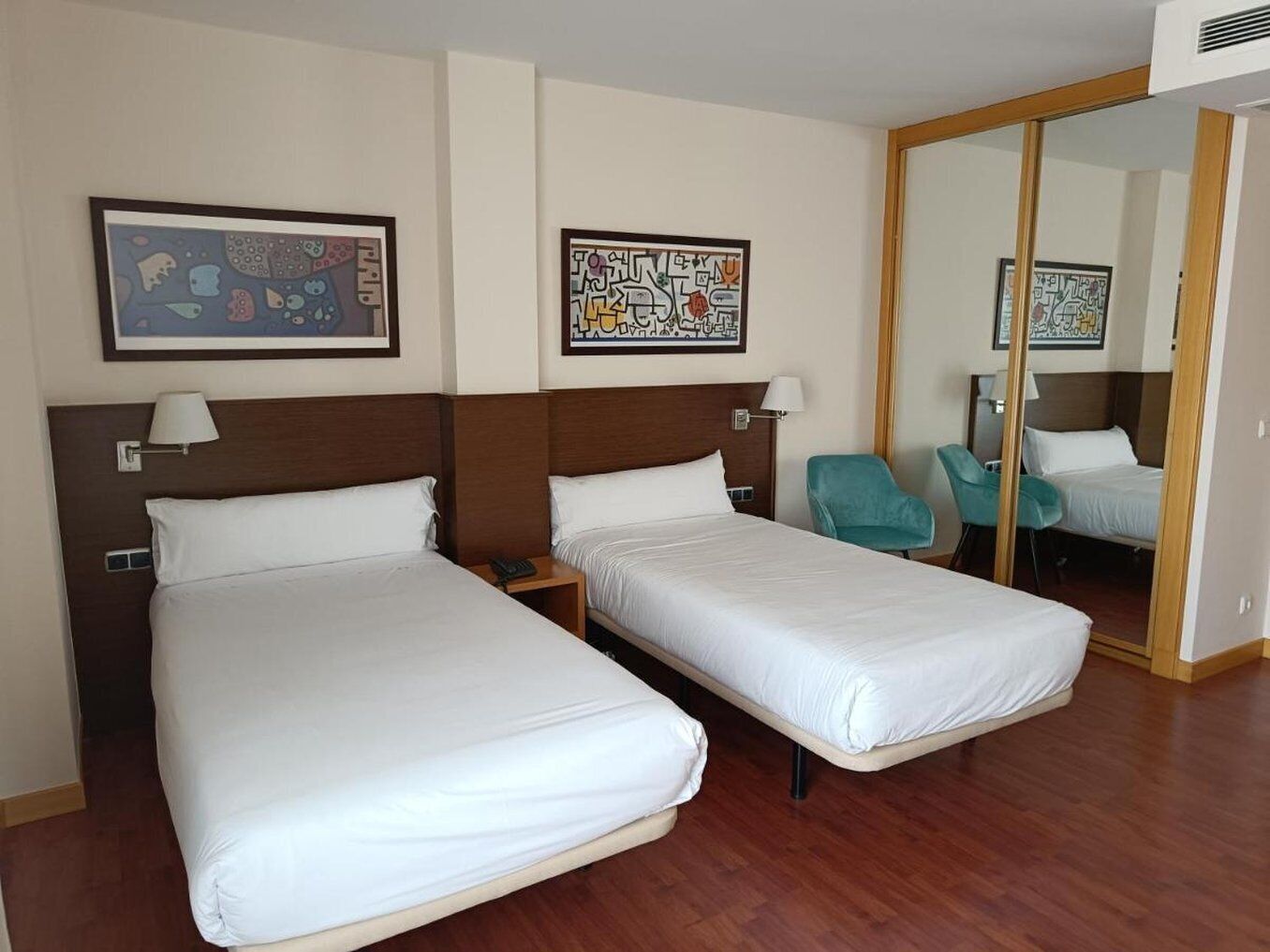Habitación de hotel con dos camas individuales, suelo de madera, un espejo grande y dos sillas.