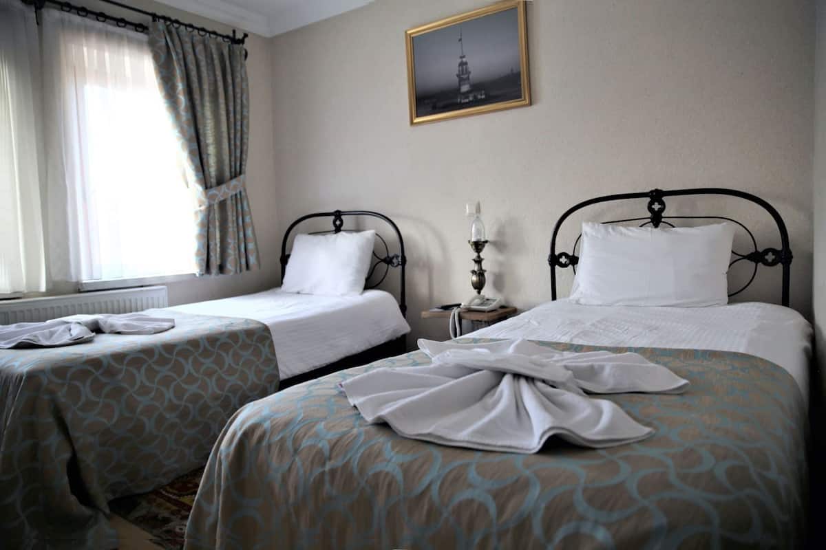 Quarto de hotel com duas camas, uma janela com cortinas, uma mesinha de cabeceira com abajur e um quadro na parede.