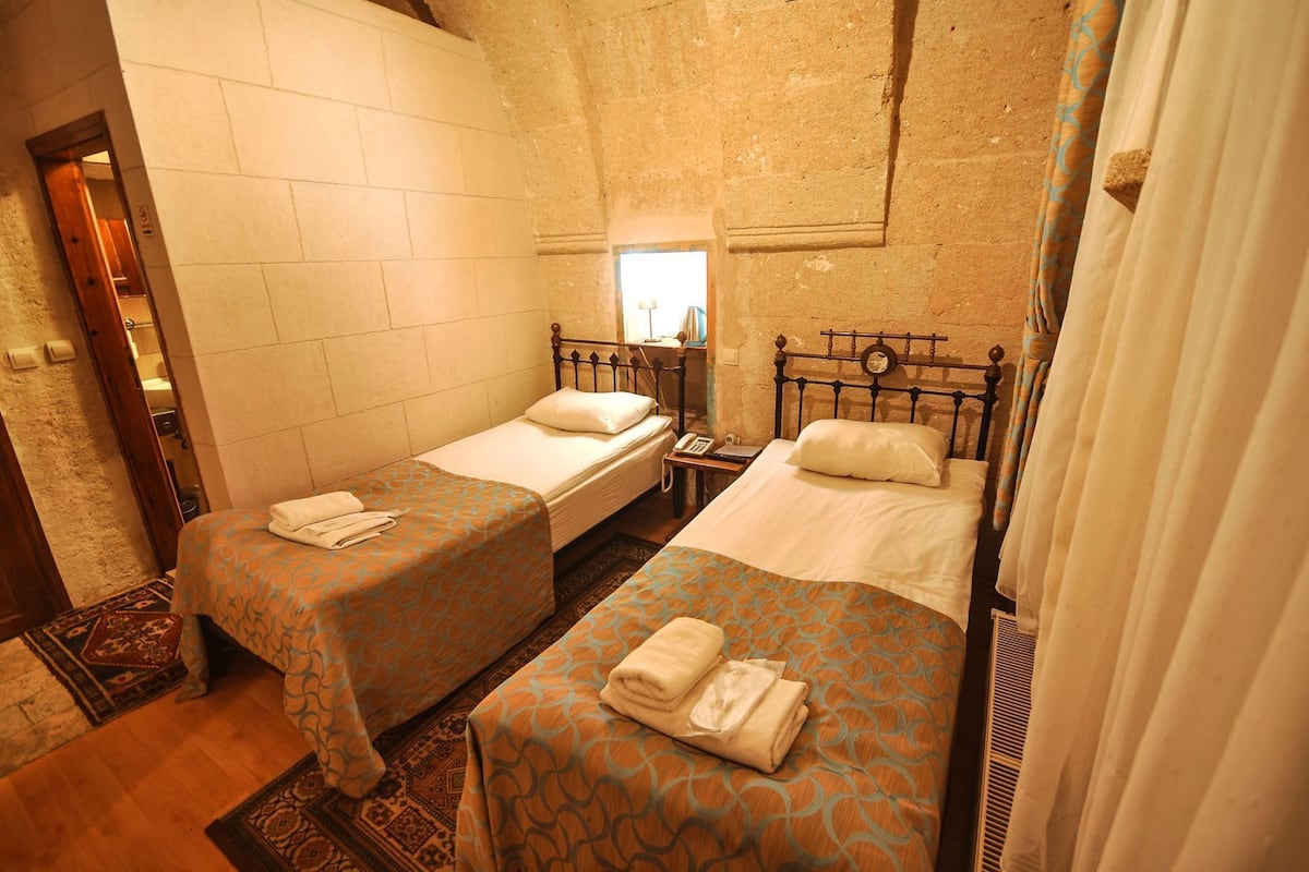 Quarto com duas camas individuais, uma mesinha com telefone e uma janela com cortinas.