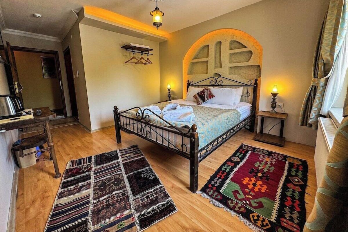 Um quarto com cama, duas mesinhas de cabeceira, luminária pendente e janela com cortinas.