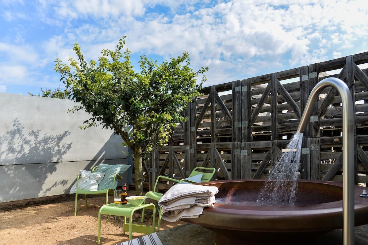 An outdoor seating area with a metal chair, a table with a bottle and a bowl, and a large metal basin with water flowing from a faucet.