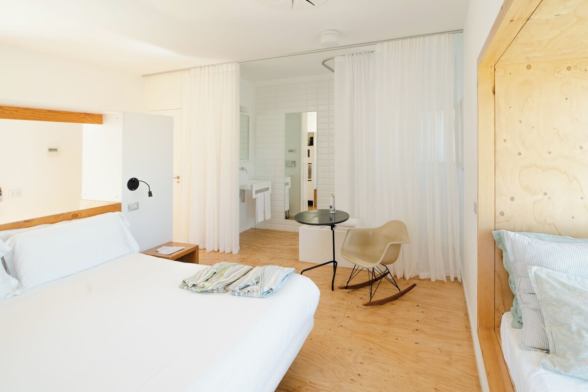 Habitación de hotel con cama, una mesita, un sillón mecedor y vistas al baño.