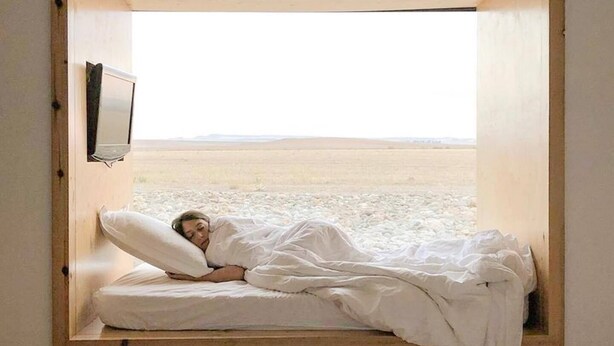 Een bed in een kamer met een groot raam, een televisie aan de muur en uitzicht op een kaal landschap.