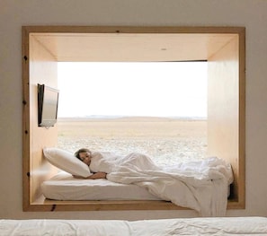 Un lit dans une chambre dotée d’une grande fenêtre, d’un téléviseur fixé au mur et offrant une vue sur un paysage désolé.