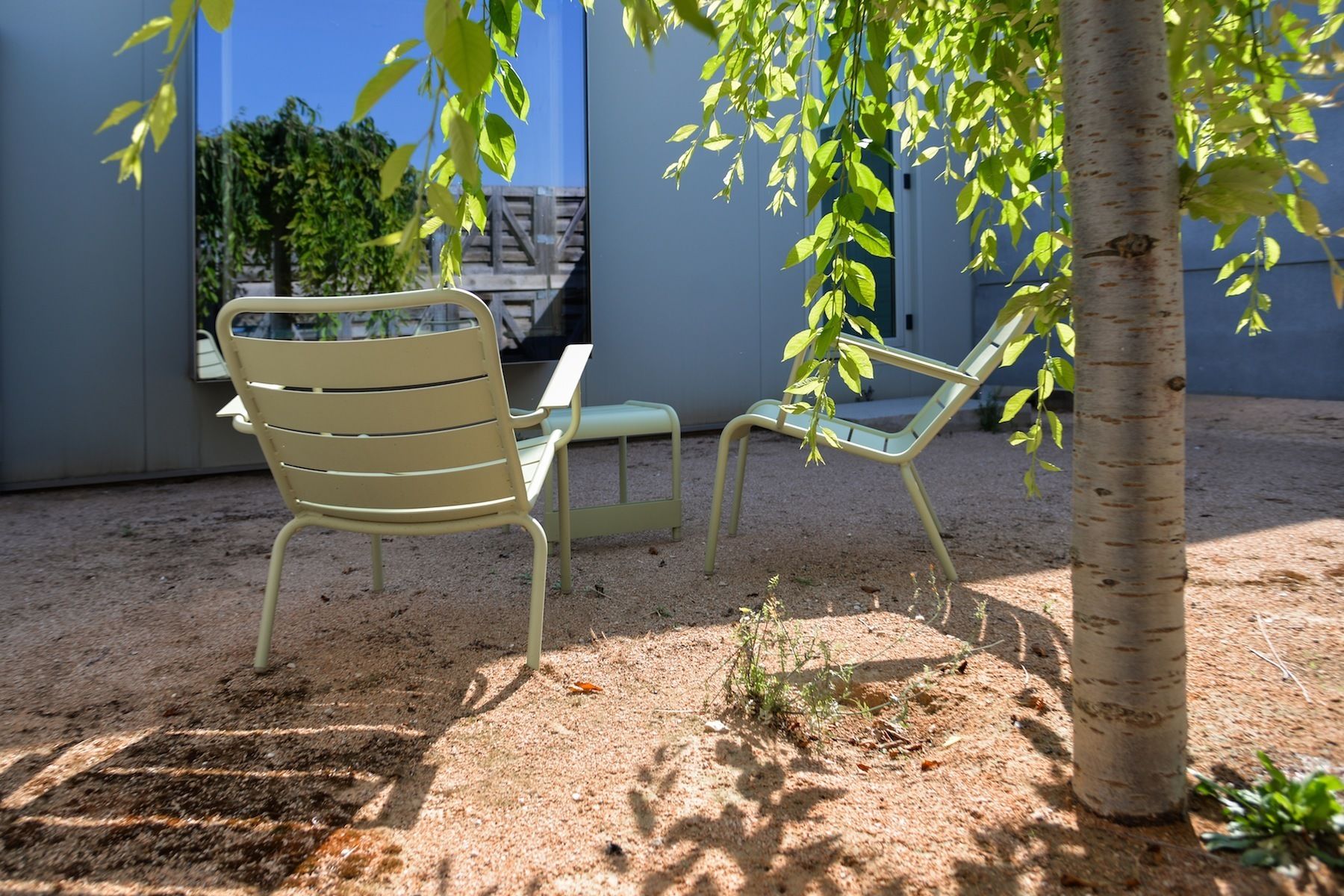 Two outdoor chairs with metal frames and green cushions, positioned on a sandy ground with a tree and a building in the background.