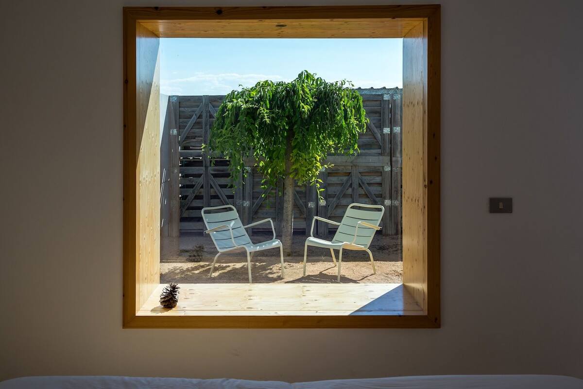 A bedroom with a large window overlooking a patio area with two chairs and a tree.