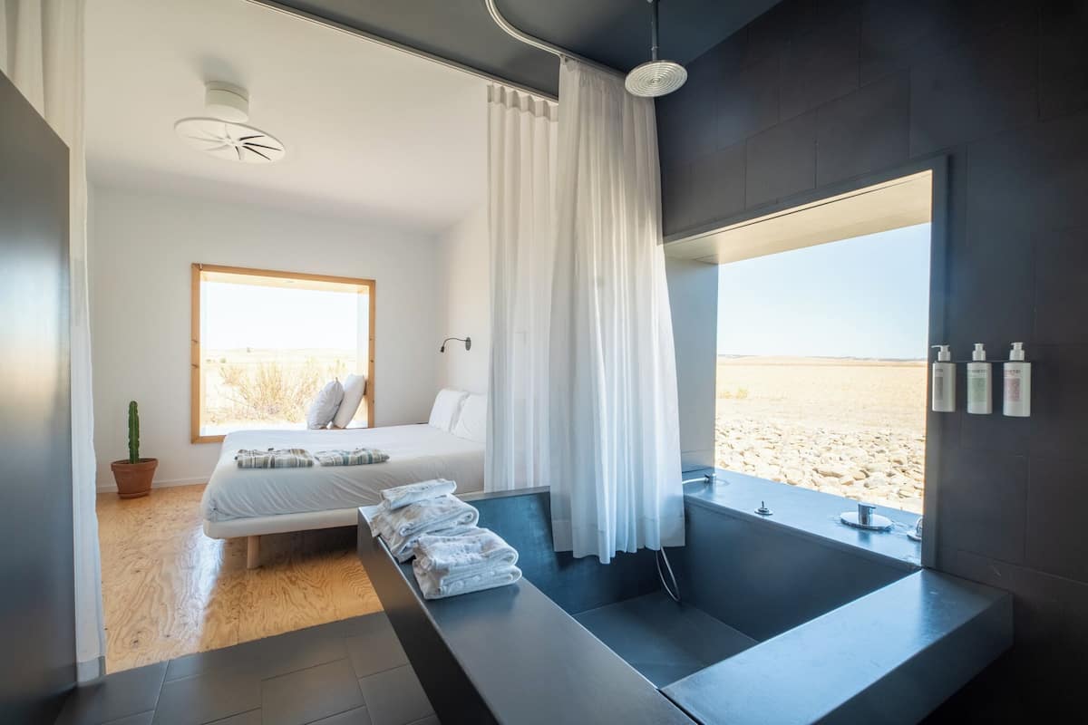 A modern bedroom with a large bed, a view of the beach, a bathtub, and a potted plant.