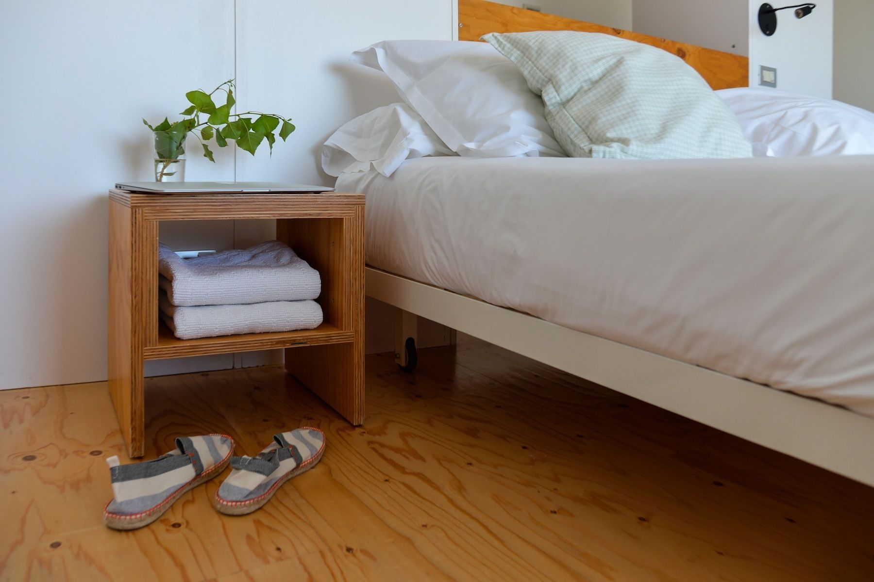 Ein ordentlich bezogenes Bett mit weißer Bettwäsche, ein Nachttisch aus Holz mit gefalteten Handtüchern, ein kleiner Topfpflanze und ein Paar Hausschuhe auf dem Boden.