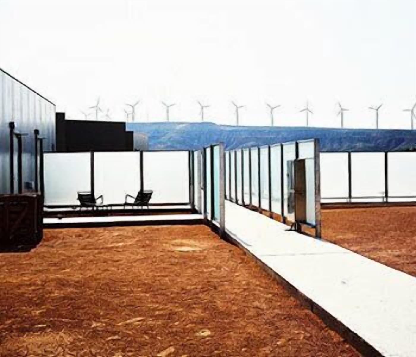 Un edificio con un pasillo largo y estrecho que conduce a una puerta de cristal, rodeado por una terraza de madera y una vista de aerogeneradores a lo lejos.