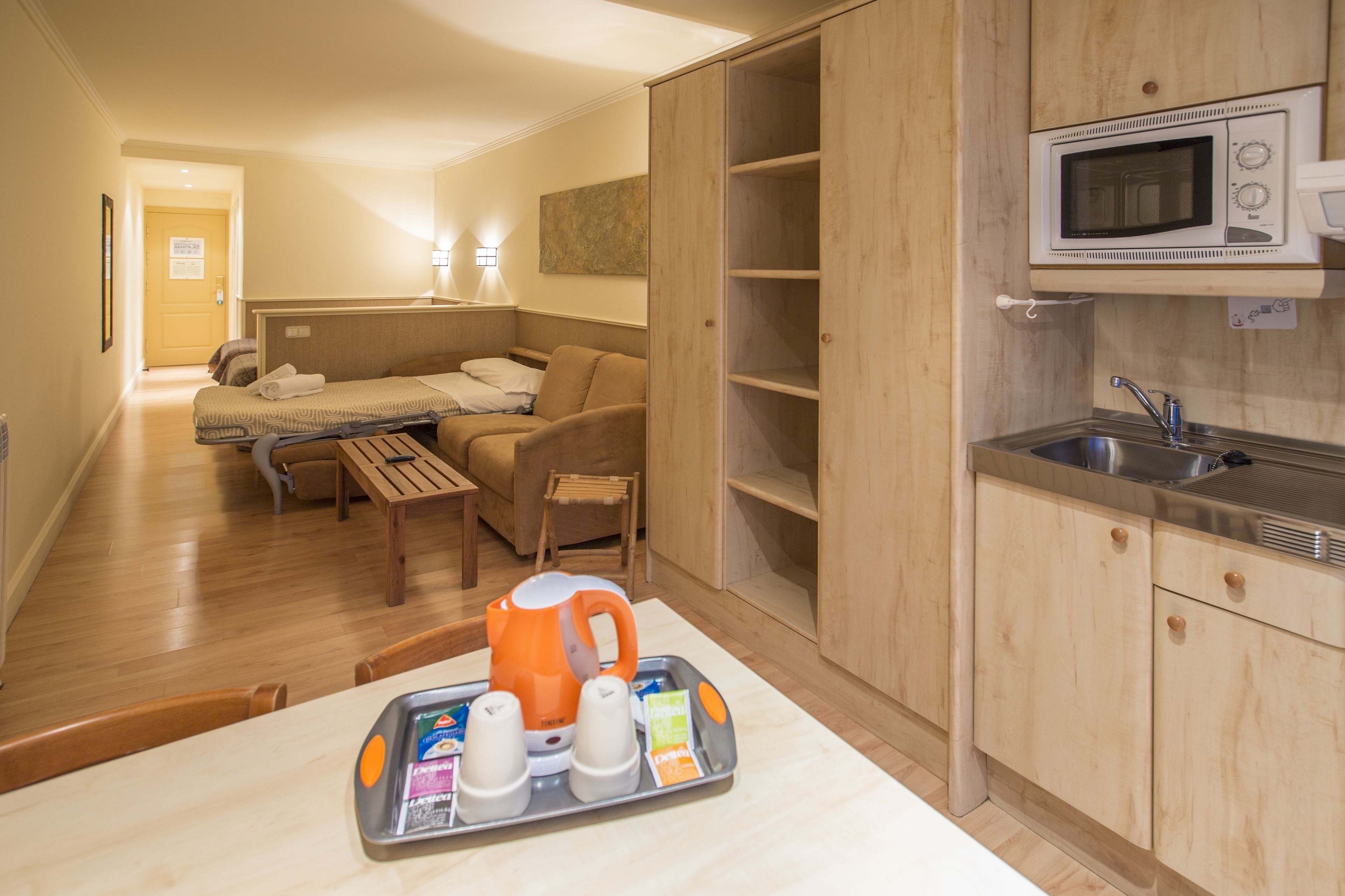 A compact hotel room with a kitchenette, a bed, a sofa, and a dining table with a tray of breakfast items.