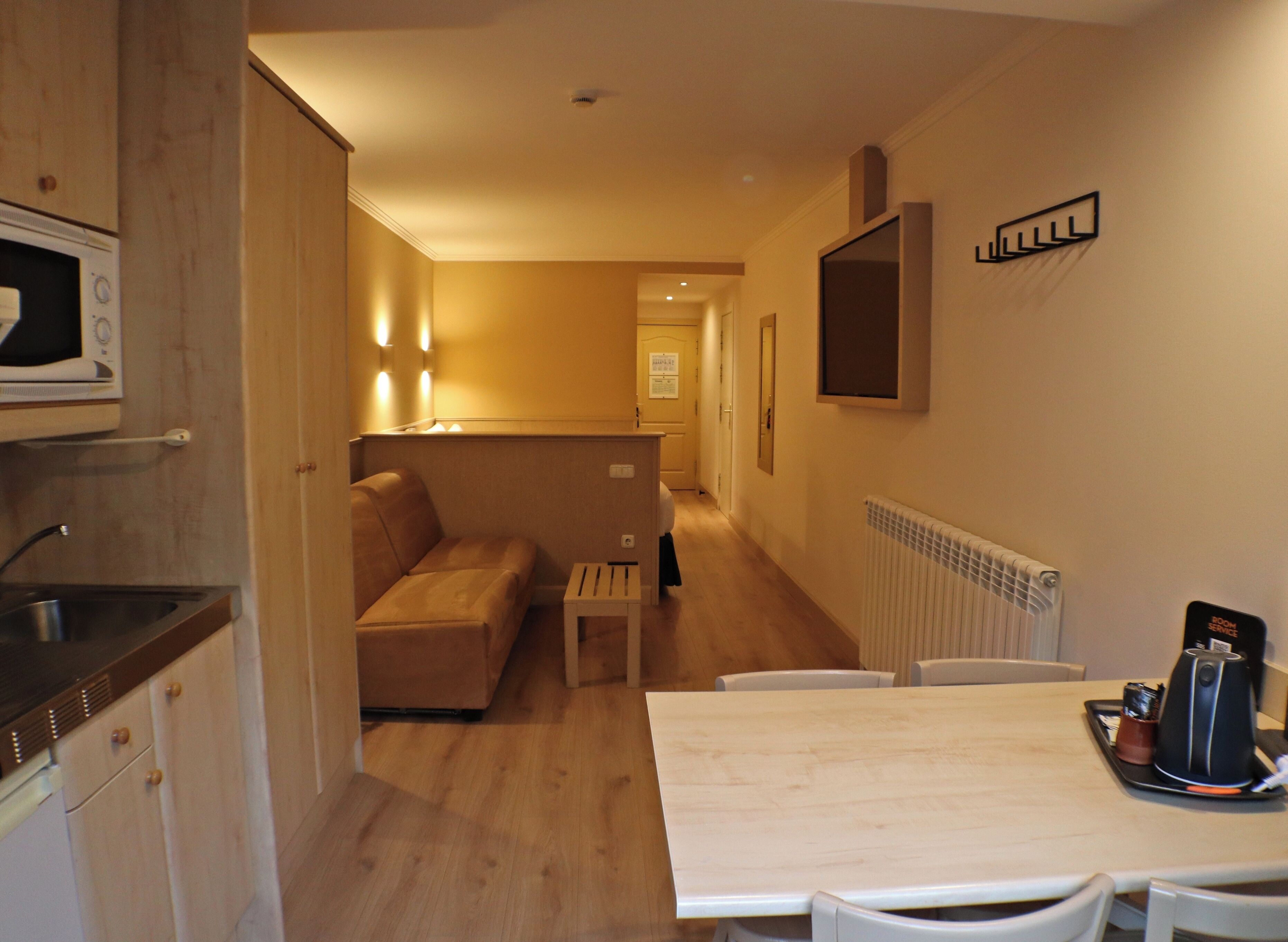A compact hotel room with a kitchenette, a sofa, a dining table, and a small coffee table.