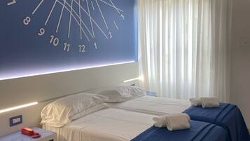 A hotel room with a large wall clock, two beds with blue bedspreads, white bedding, a bedside table with a red telephone, and a patterned floor.