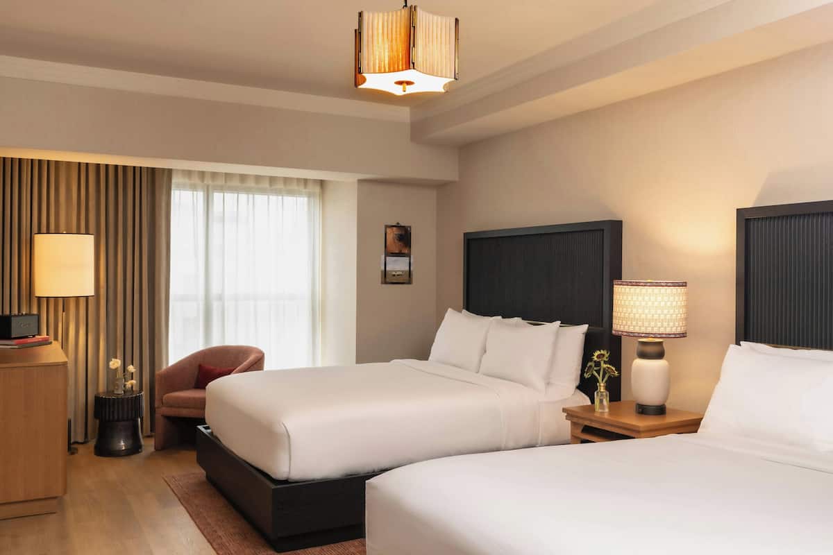 A hotel room with two beds, a desk, a chair, a lamp, and a window with curtains.