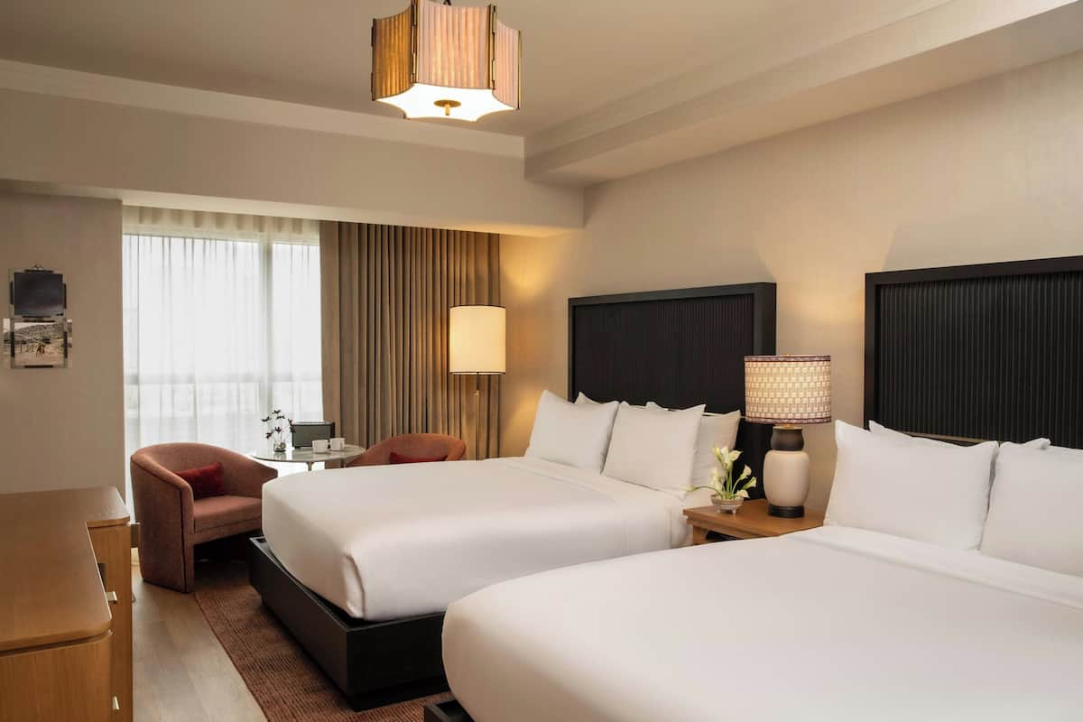 A hotel room with two beds, a desk, a chair, a lamp, and a view of the outdoors.