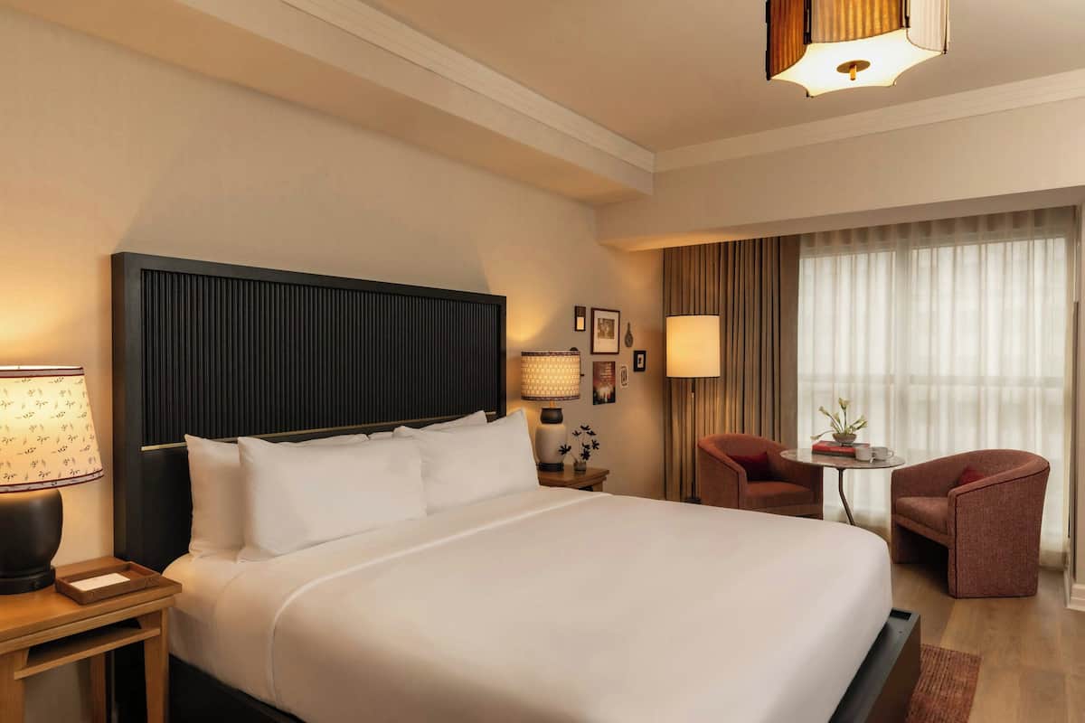 A hotel room with a large bed, bedside tables, a desk, a chair, and a window with curtains.