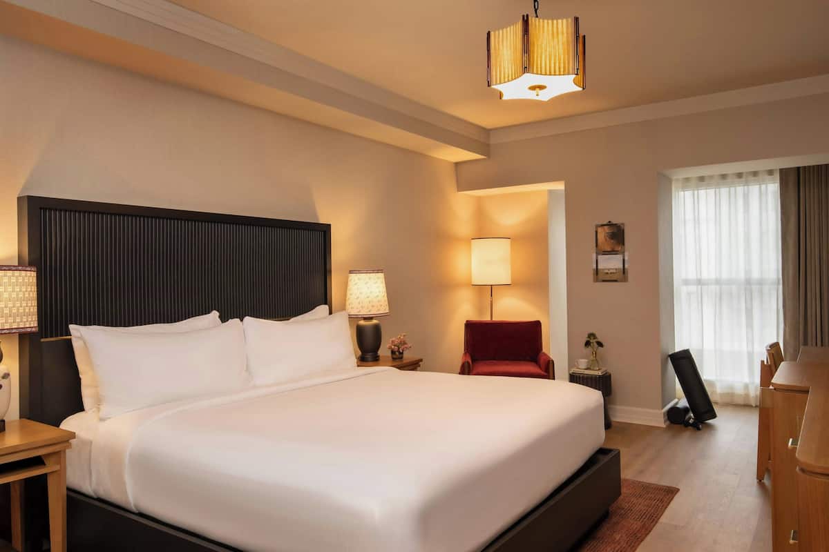 A hotel room with a large bed, bedside lamps, a red chair, a desk, and a large window with curtains.
