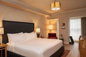 A hotel room with a large bed, bedside lamps, a red chair, a desk, and a large window with curtains.