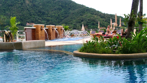 A pool area with a waterfall feature, lounge chairs, and a view of a forested hill. - Peach Hill Resort (Karon)