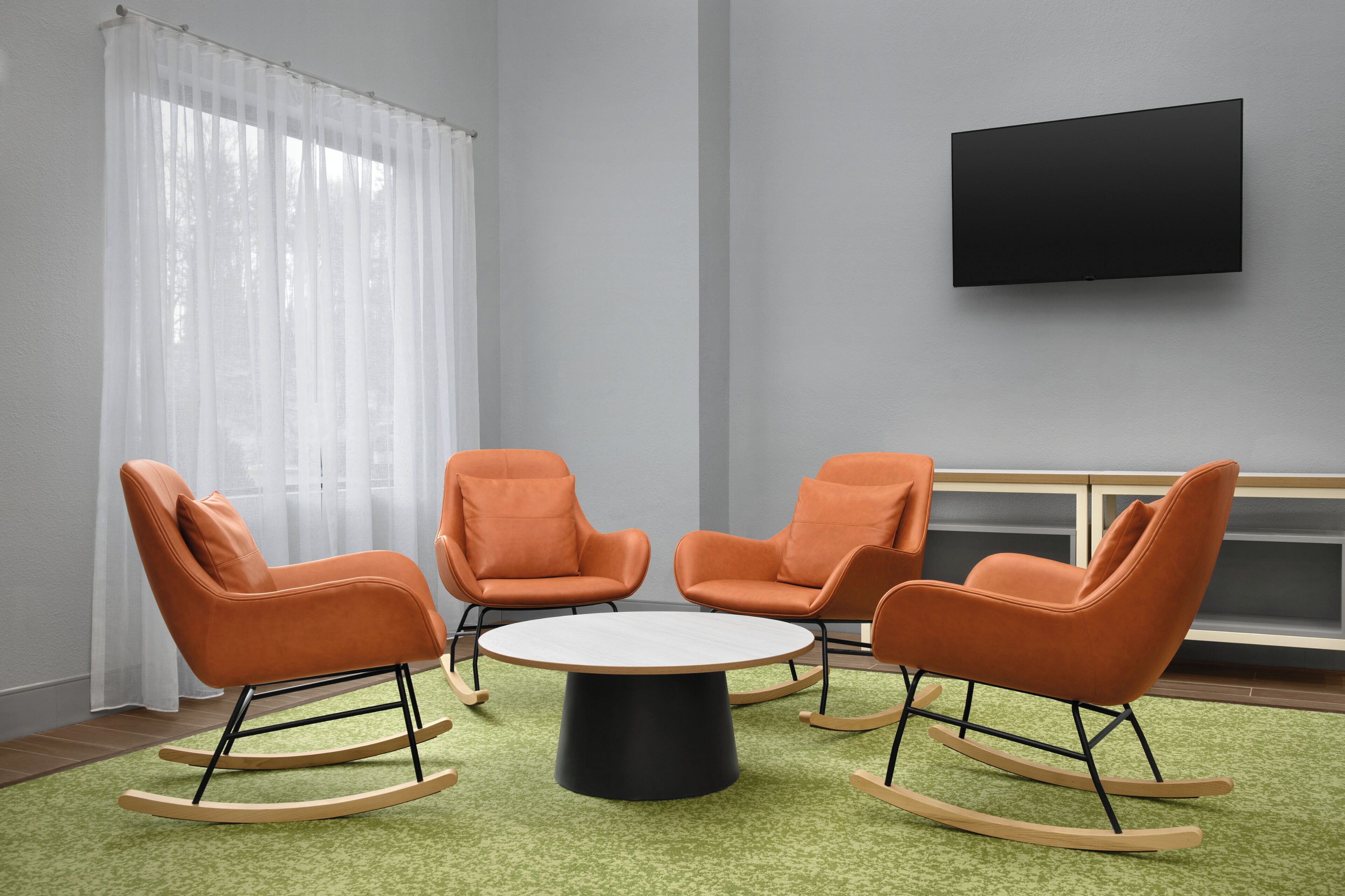 Ein Zimmer mit einem runden Tisch, drei orangefarbenen Schaukelstühlen, einem Flachbildfernseher an der Wand und einem Fenster mit durchscheinenden Vorhängen.