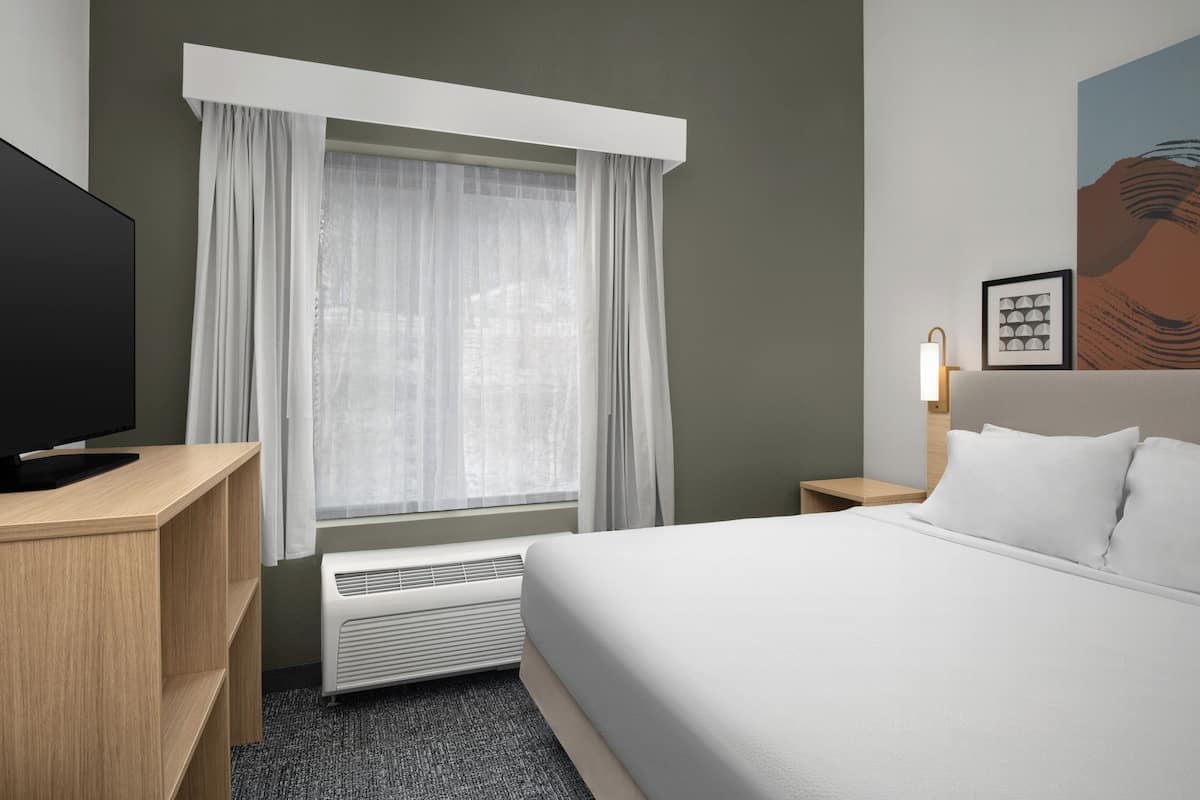 Habitación de hotel con cama, televisión, calefacción, mesita de noche, lámpara y ventana con cortinas.
