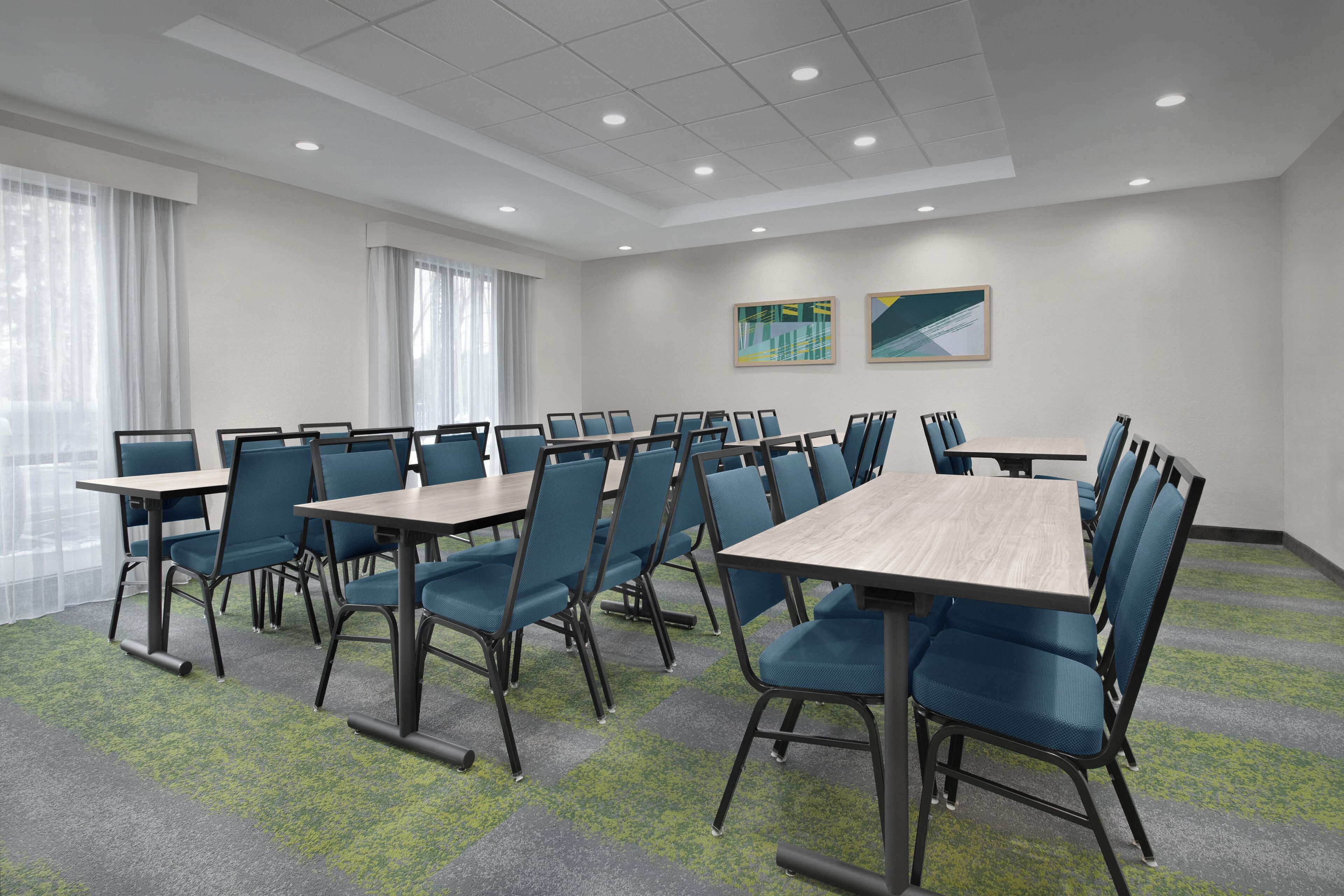 A conference room with tables and chairs, large windows with curtains, and two abstract paintings on the wall.