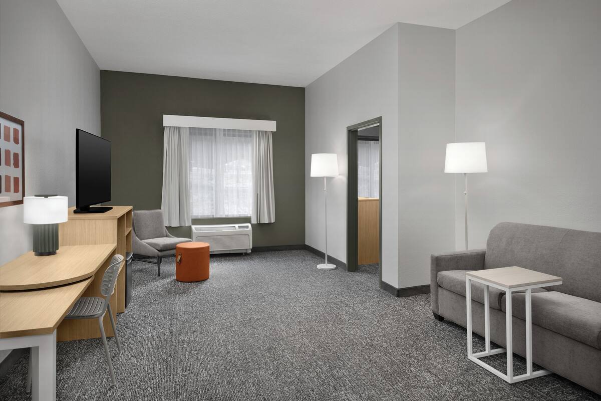 Habitación de hotel con escritorio, silla, sofá, dos lámparas, televisión y ventana con cortinas.