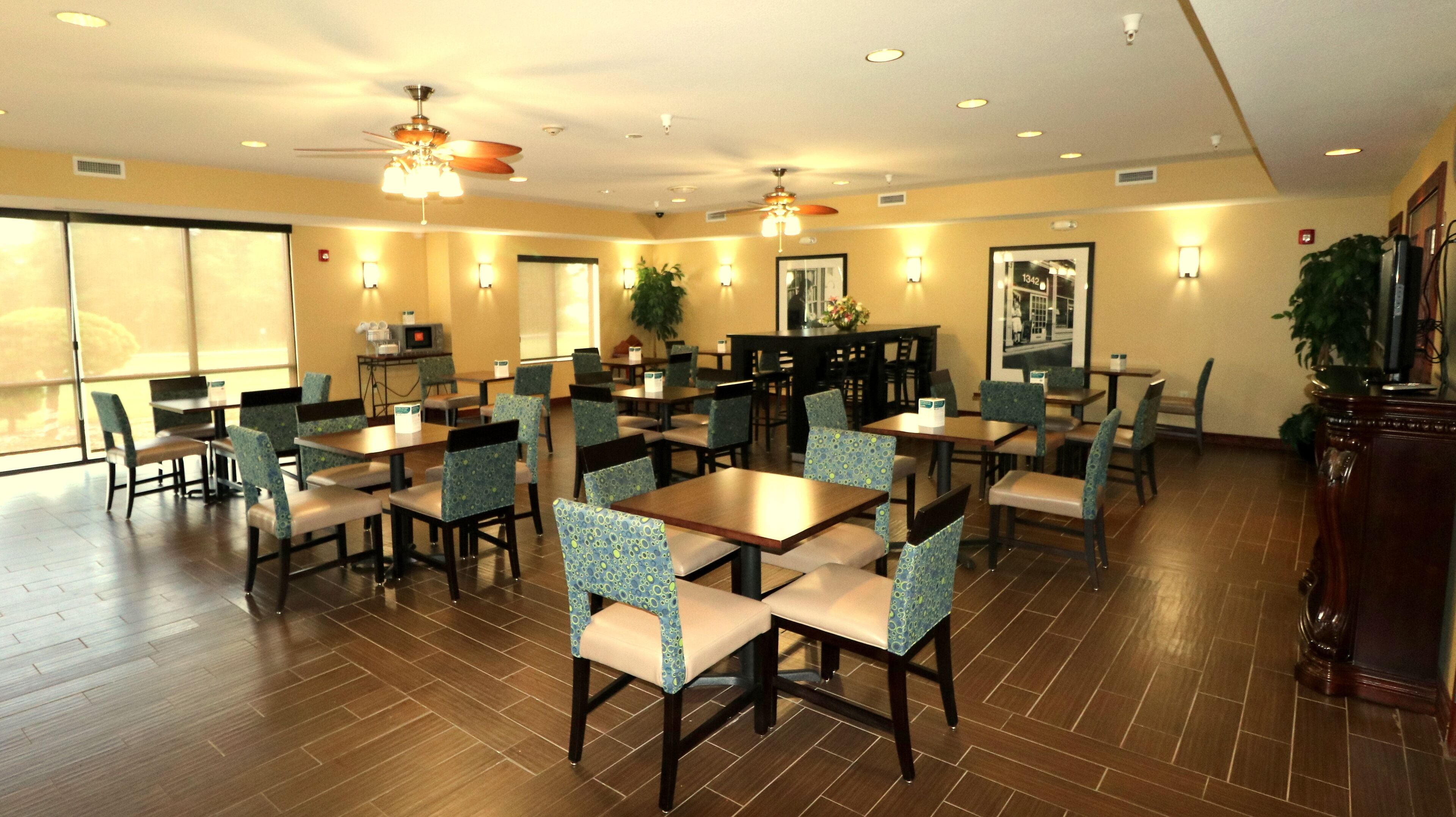 A hotel dining area with multiple tables and chairs, ceiling fans, and wall-mounted lights.