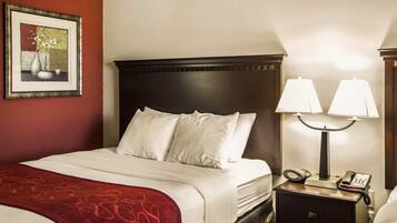 A neatly made bed with white linens, a red throw blanket, and a dark wooden headboard. There is a nightstand with two lamps and a telephone. A framed picture hangs on the wall to the left.