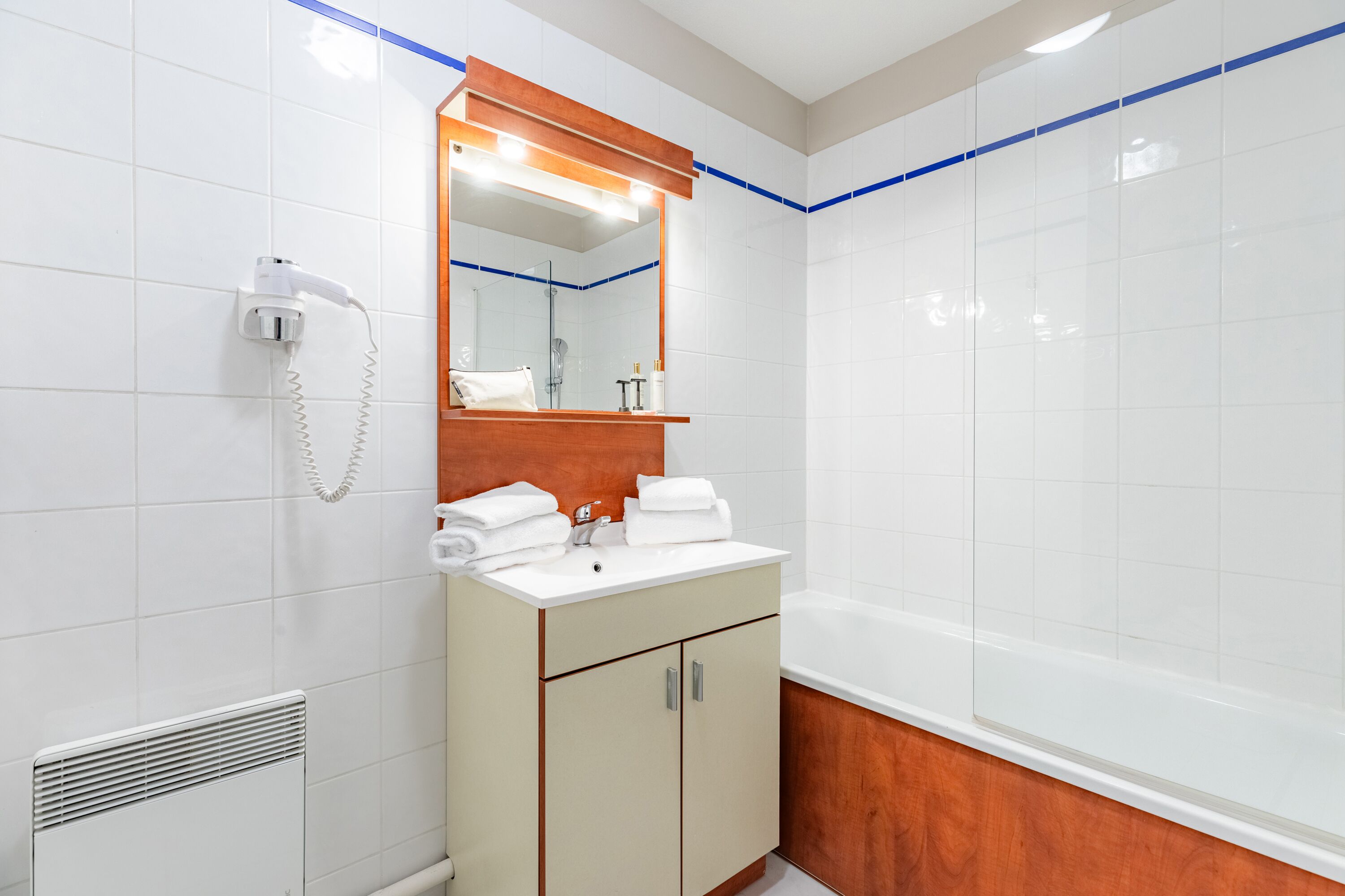 Un baño con azulejos blancos, un tocador de madera, un espejo, un lavabo blanco, una bañera y un secador de pelo.
