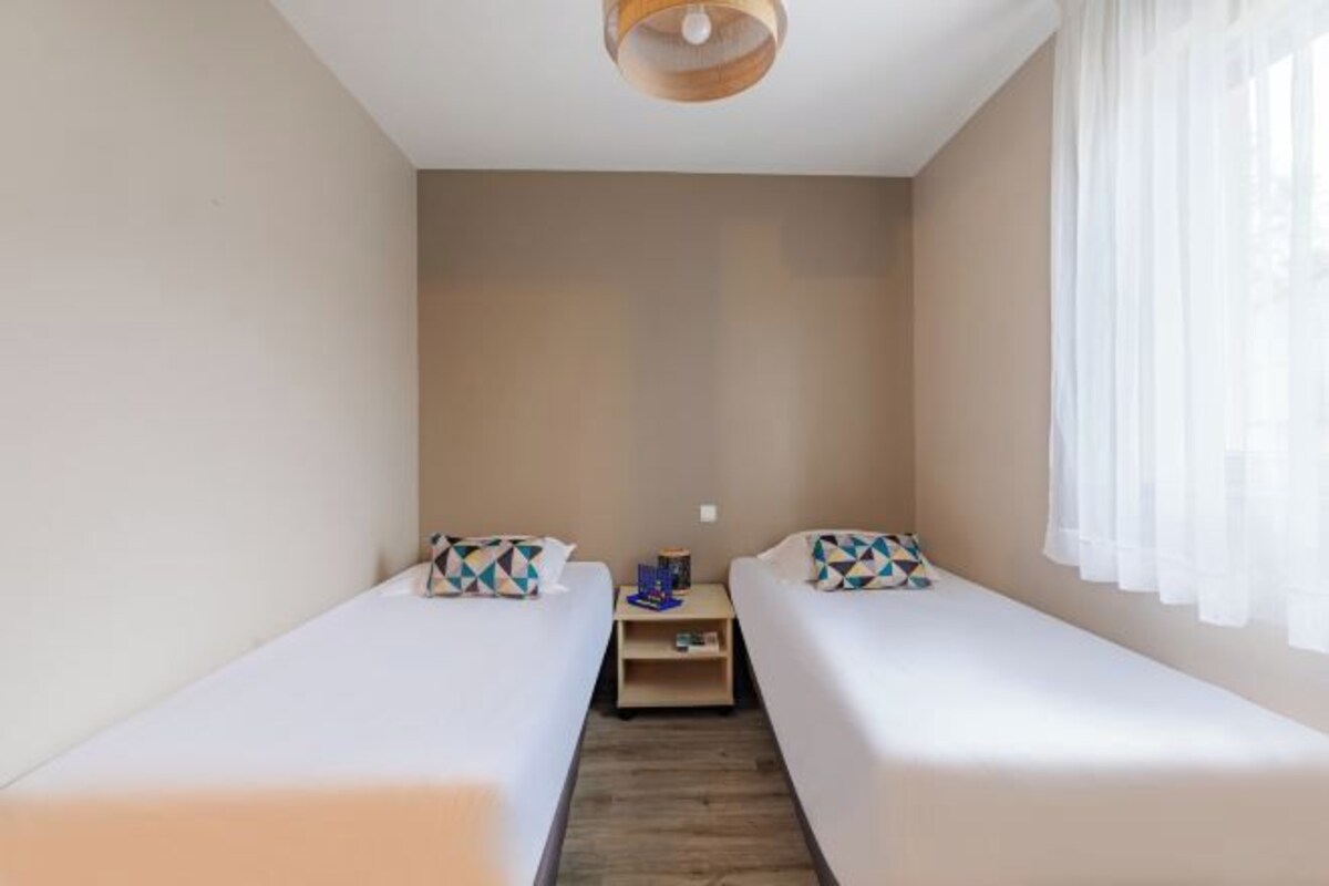 A small, single-room accommodation with two single beds, a wooden floor, a small table, and a window with sheer curtains.