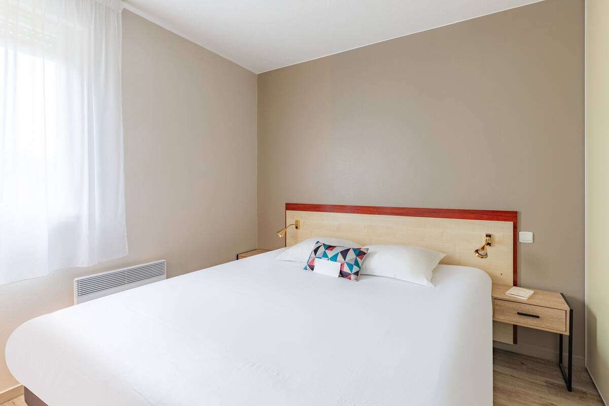 Une chambre d’hôtel avec un grand lit, une table de chevet, une commode et une fenêtre avec des rideaux.