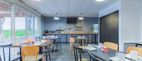 Una cafetería moderna con mesas, sillas, una zona de cocina y grandes ventanales con vistas.