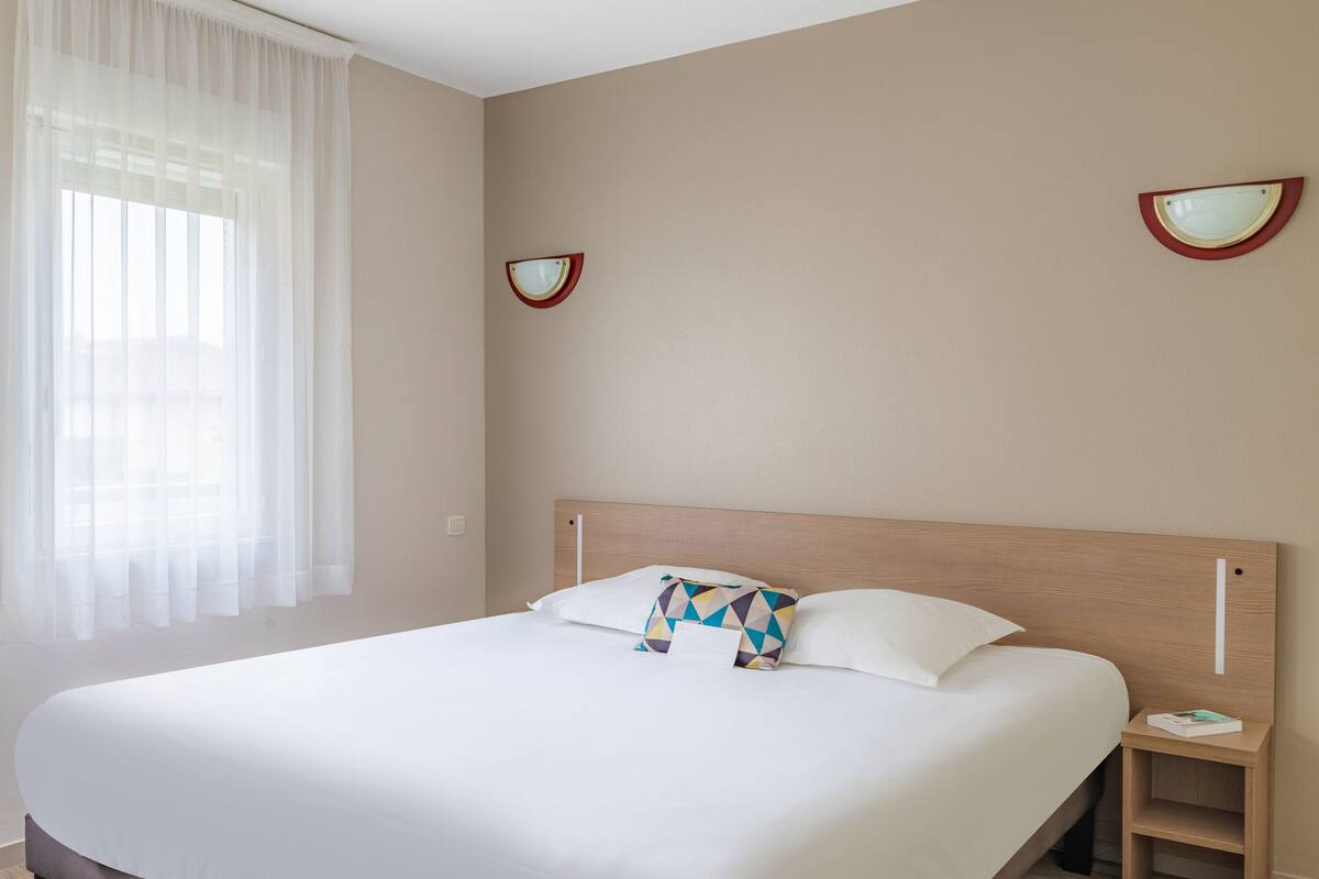 Un lit bien fait, avec du linge de lit blanc et un coussin à motifs géométriques, une table de chevet avec un livre, et un luminaire fixé au mur.