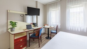 Habitación de hotel con cama, escritorio con computadora portátil, silla, pequeña mesa con frutas, planta en maceta y televisor montado en la pared.