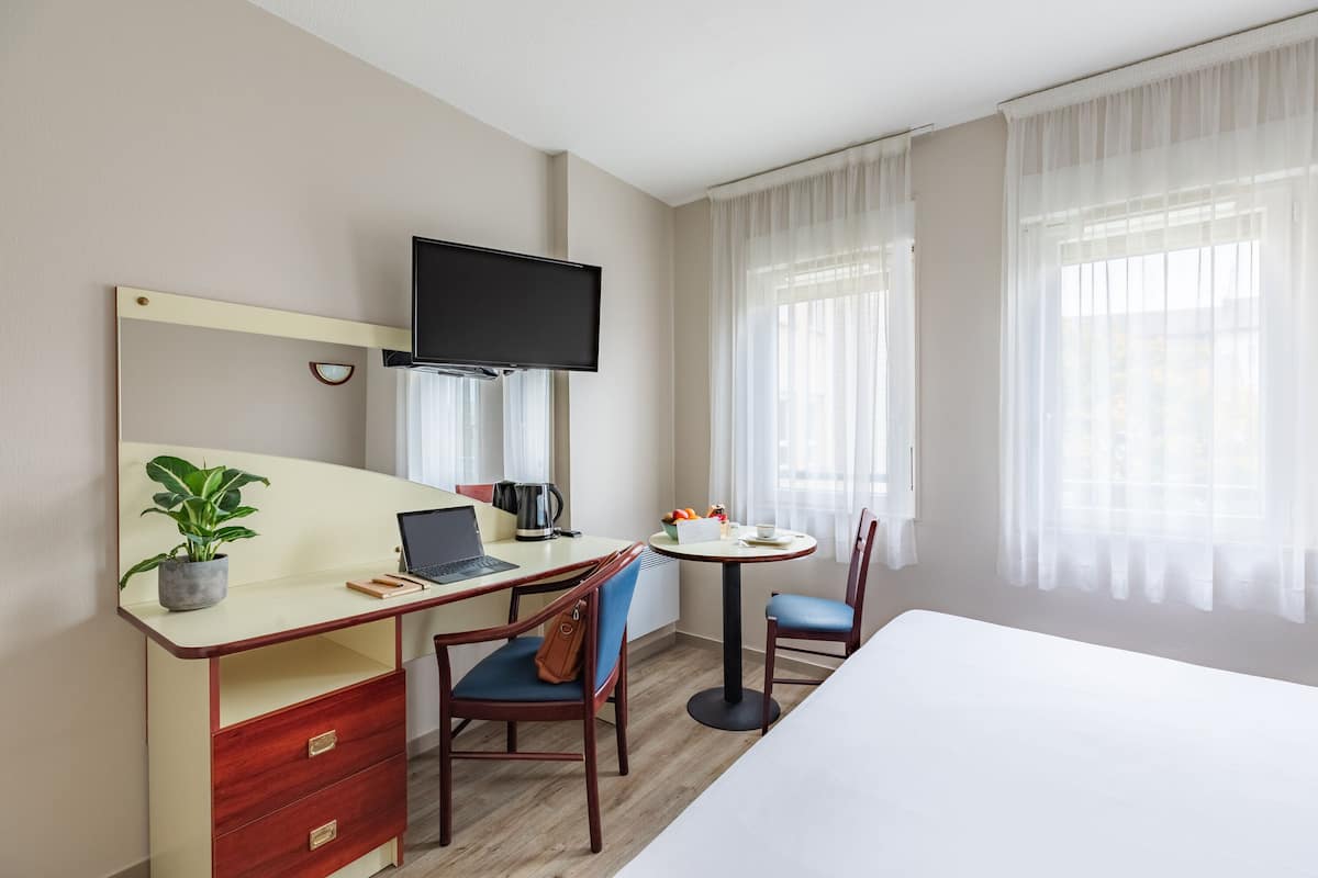 Une chambre d’hôtel avec un lit, un bureau avec un ordinateur portable, une chaise, une petite table avec des fruits, une plante en pot et un téléviseur fixé au mur.
