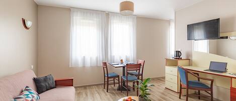A modern, well-lit room with a sofa, dining table, chairs, a desk with a laptop, a TV, and a plant on a small table.