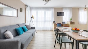 Una sala moderna con un sofá gris, cojines color turquesa, una mesa de comedor con sillas, un ventilador de techo y un televisor en la pared.