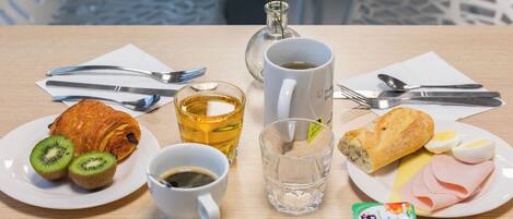 Una mesa de madera con bollos de croissant, rebanadas de kiwi, café, té, pan, huevos, jamón y un recipiente de yogur.