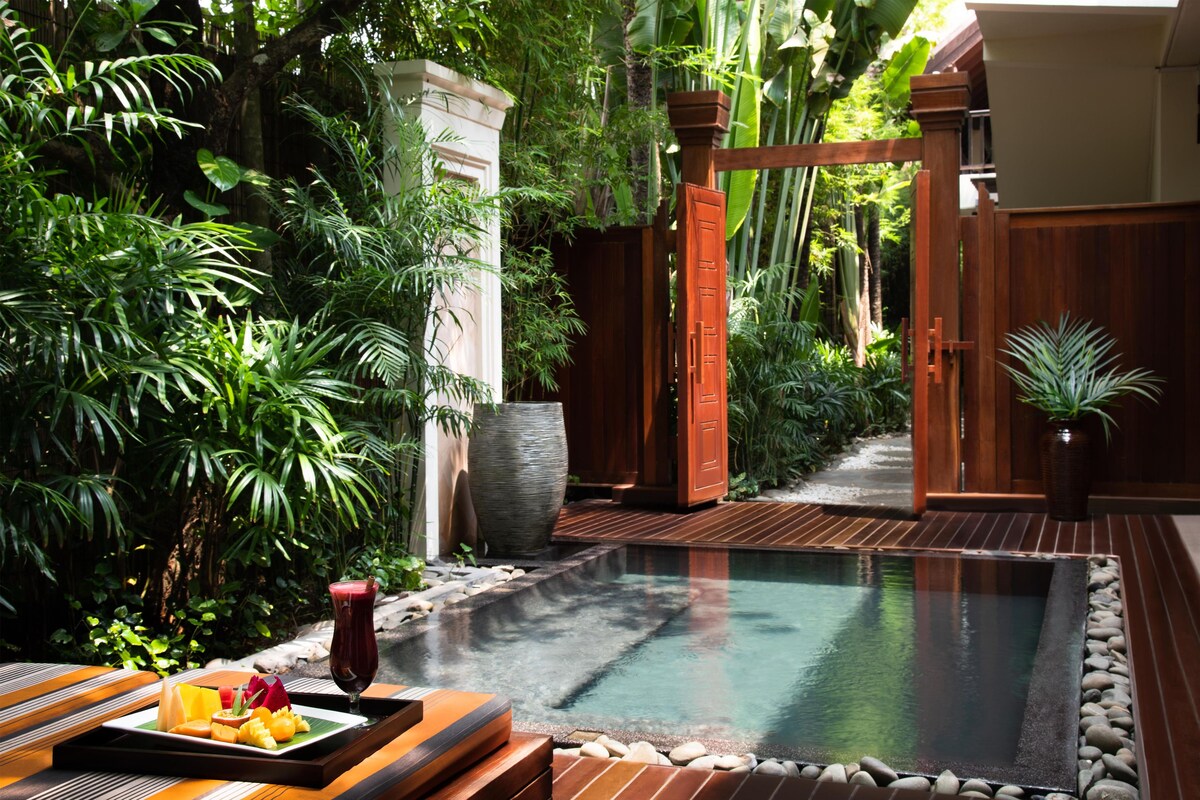 A serene pool area with a wooden deck, surrounded by lush greenery and a stone pathway.