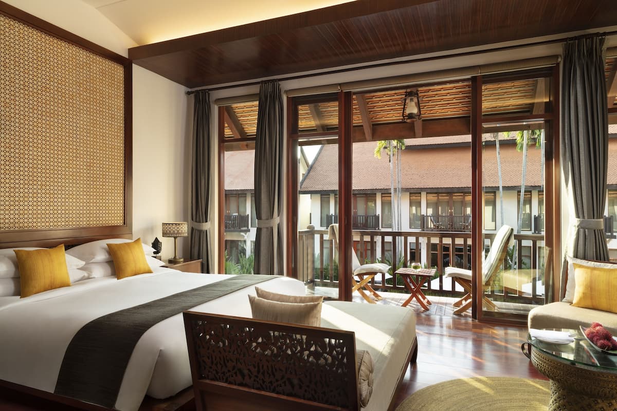 A hotel room with a large bed, a sitting area with a table and chairs, and a balcony with a view of buildings.